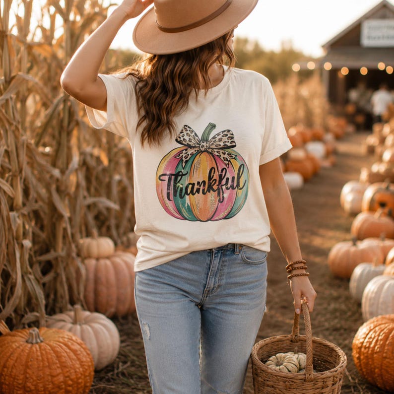 Woman walking through a pumpkin patch wearing a colorful Thankful pumpkin t-shirt with a stylish wide-brim hat and basket.