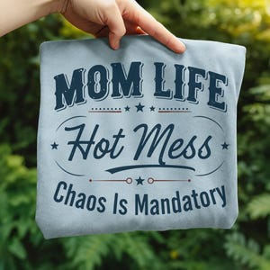 May include: A light blue t-shirt with the words "MOM LIFE Hot Mess Chaos Is Mandatory" in navy blue lettering. The design includes stars and decorative lines. The shirt is held up against a green background.