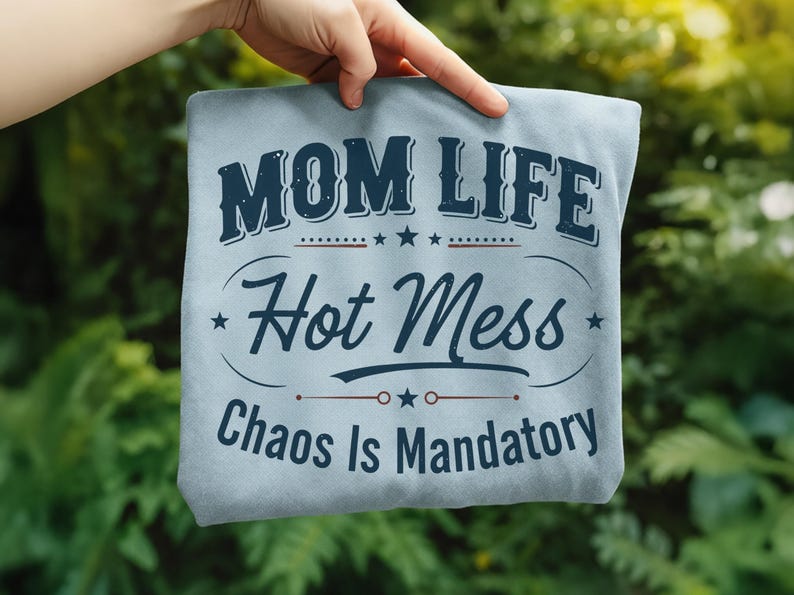 May include: A light blue t-shirt with the words "MOM LIFE Hot Mess Chaos Is Mandatory" in navy blue lettering. The design includes stars and decorative lines. The shirt is held up against a green background.