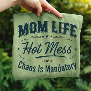 May include: A light green t-shirt with the text "MOM LIFE Hot Mess Chaos Is Mandatory" in navy blue. The text is arranged in a vintage style with stars and decorative lines. The shirt is held up by a hand against a blurred green background.