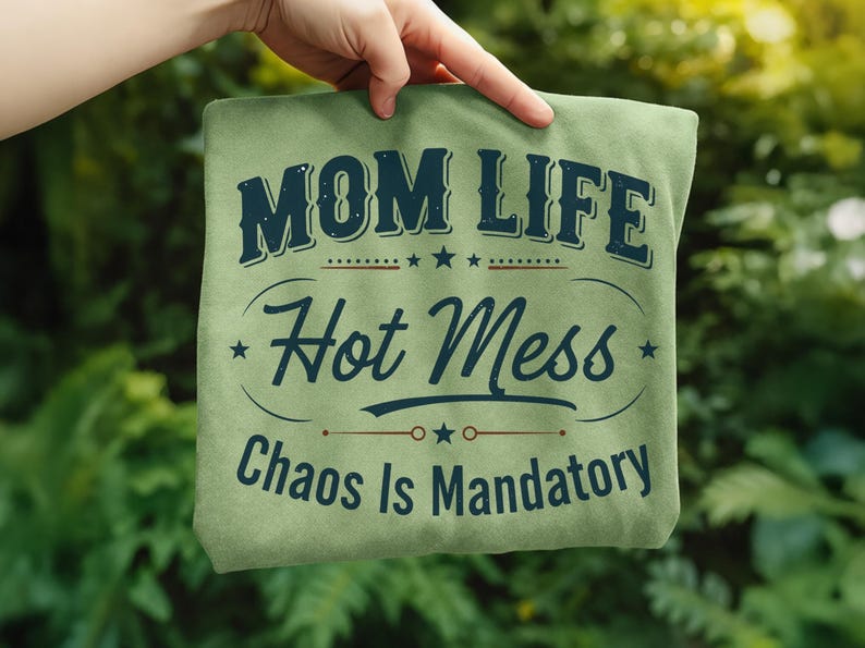 May include: A light green t-shirt with the text "MOM LIFE Hot Mess Chaos Is Mandatory" in navy blue. The text is arranged in a vintage style with stars and decorative lines. The shirt is held up by a hand against a blurred green background.