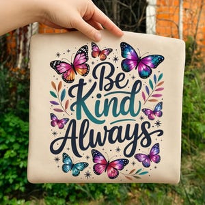 May include: A beige tote bag featuring colorful butterflies and the words "Be Kind Always" in a whimsical font. The butterflies are in shades of pink, purple, orange, and blue. The bag is held up by a hand.