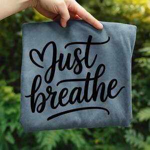 May include: A slate blue t-shirt with the words "Just breathe" in a black, cursive font. The word "Just" is above "breathe", with a heart shape replacing the letter "u". The shirt is held up against a green, leafy background.