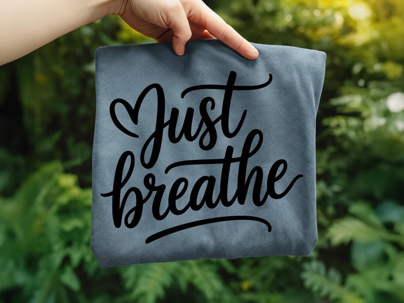 May include: A slate blue t-shirt with the words "Just breathe" in a black, cursive font. The word "Just" is above "breathe", with a heart shape replacing the letter "u". The shirt is held up against a green, leafy background.