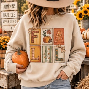 Op de afbeelding: Een beige sweater met ronde hals en een grafische print van zes rechthoekige panelen in twee rijen. De panelen bevatten de letters F, A, L, L, samen met illustraties van een pompoen, een eikel, bloemen en decoratieve strikken.