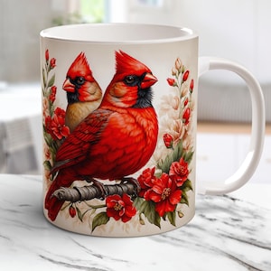 May include: White ceramic mug featuring a detailed illustration of two red cardinal birds perched on a branch, surrounded by red flowers and foliage. The mug has a white handle and a cream-colored background.
