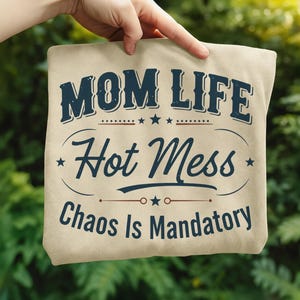May include: A beige tote bag with the text "MOM LIFE Hot Mess Chaos Is Mandatory" in navy blue. The text is arranged in a vintage style with stars and decorative lines. The bag is held up by a hand.