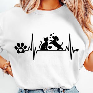 以下が含まれることがあります： 猫と犬、ハート、心拍ラインの黒いグラフィックデザインが施された白いTシャツ。左側にはハートの足跡があります。このデザインはペットと動物愛を祝い、動物好きに最適です。