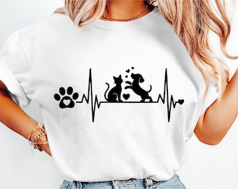 Puede incluir: Camiseta blanca con un diseño gráfico negro de un gato y un perro con corazones, sobre una línea de latidos. Una huella de pata con un corazón está a la izquierda. El diseño celebra mascotas y el amor por los animales.
