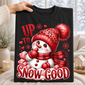 Black t-shirt displaying a red and black snowman winter design with Up to Snow Good typography, snowflakes and plaid scarf