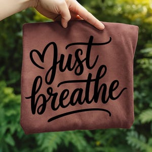 May include: A maroon-colored t-shirt with the words "Just breathe" in a black, cursive font. The shirt is held up against a backdrop of green foliage.