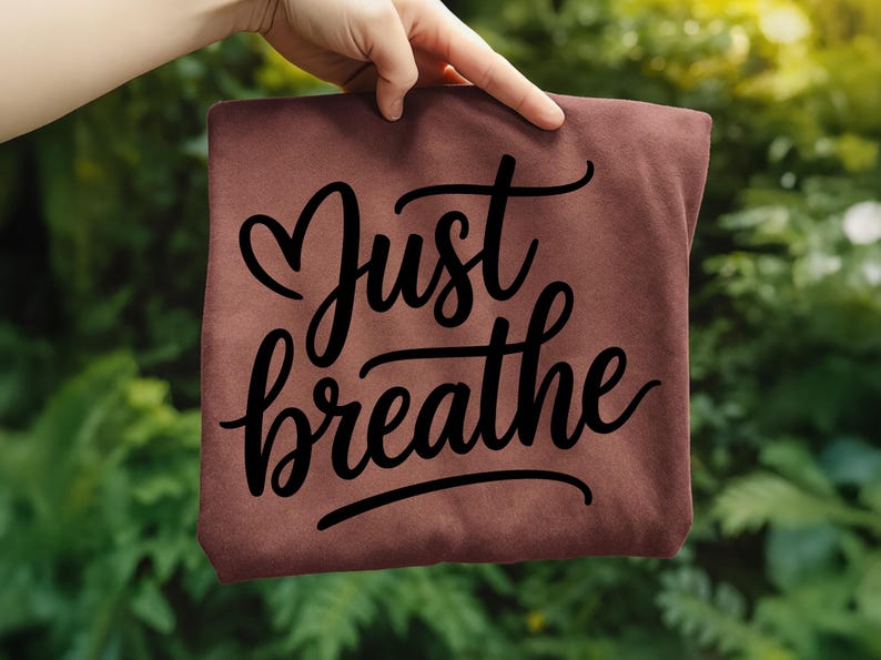 May include: A maroon-colored t-shirt with the words "Just breathe" in a black, cursive font. The shirt is held up against a backdrop of green foliage.