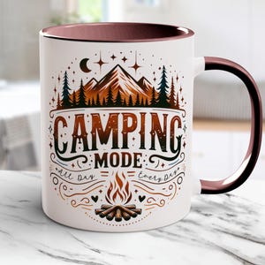 Könnte beinhalten: Weißer Keramikbecher mit braunem Henkel und Innenraum. Der Becher zeigt die Worte "CAMPING MODE" in einer dekorativen Schriftart, zusammen mit Illustrationen von Bergen, Bäumen, einem Lagerfeuer und dem Text "all day, everyday."