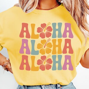 Pode incluir: Camiseta amarela com a palavra "ALOHA" repetida tr&ecirc;s vezes em letras de estilo retr&ocirc;. Cada "ALOHA" tem uma cor diferente, com um gr&aacute;fico de flor de hibisco no centro de cada palavra.