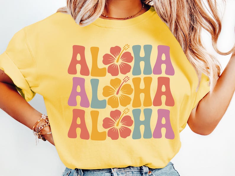 Pode incluir: Camiseta amarela com a palavra "ALOHA" repetida tr&ecirc;s vezes em letras de estilo retr&ocirc;. Cada "ALOHA" tem uma cor diferente, com um gr&aacute;fico de flor de hibisco no centro de cada palavra.