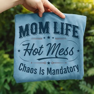 May include: A light blue t-shirt with the words "MOM LIFE Hot Mess Chaos Is Mandatory" in navy blue lettering. The shirt is held up against a green, leafy background.