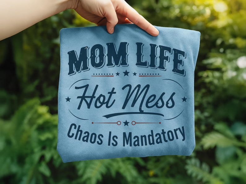 May include: A light blue t-shirt with the words "MOM LIFE Hot Mess Chaos Is Mandatory" in navy blue lettering. The shirt is held up against a green, leafy background.