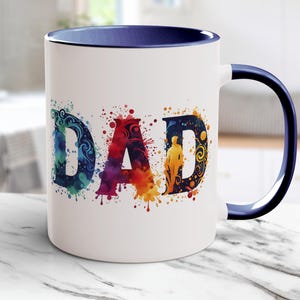 Może przedstawiać: Biały ceramiczny kubek z niebieskim wnętrzem i uchwytem. Na przodzie nadrukowano słowo "DAD" w kolorowej akwarelowej czcionce. Litery zdobią plamy koloru i elementy dekoracyjne.