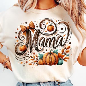 May include: Cream-colored sweatshirt with a fall-themed graphic. The design features the word "Mama" in a decorative script, surrounded by pumpkins, leaves, and stars. A brown polka-dot bow is at the top.