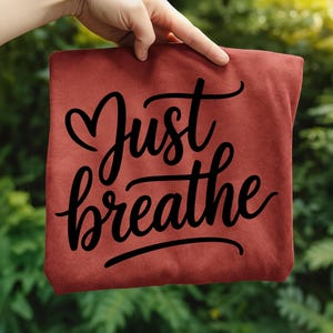 May include: A maroon-colored t-shirt with the words "Just breathe" in a black, cursive font. The phrase is centered on the shirt, with the word "Just" above "breathe". The shirt is held up against a blurred green background.