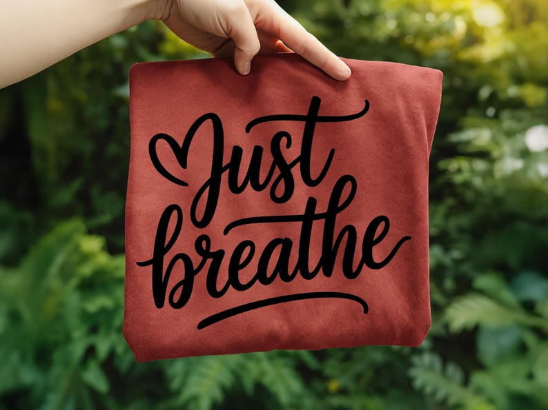 May include: A maroon-colored t-shirt with the words "Just breathe" in a black, cursive font. The phrase is centered on the shirt, with the word "Just" above "breathe". The shirt is held up against a blurred green background.