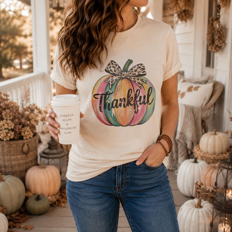 Woman wearing a cream t-shirt featuring a colorful Thankful pumpkin design with leopard bow while holding a pumpkin spice coffee on a cozy fall porch.