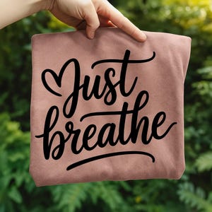 May include: A dusty rose-colored t-shirt with the words "Just breathe" in a black, cursive font. The word "Just" has a heart shape above it. The shirt is held up by a hand against a blurred green background.