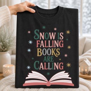 Puede incluir: Una camiseta negra de cuello redondo con el texto "SNOW IS FALLING BOOKS ARE CALLING" en letras de color verde azulado, rosa y dorado. El diseño incluye pequeños detalles en forma de estrella y un gráfico de un libro abierto con páginas rosas.