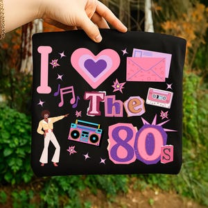 Op de afbeelding: Zwarte tote bag met een kleurrijk jaren 80 thema ontwerp. Het ontwerp bevat een roze hart, muzieknoten, een boombox en de tekst "I Love The 80s". Het ontwerp bevat ook een discodanser en sterren.