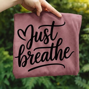 May include: A maroon-colored t-shirt with the words "Just breathe" in a black, cursive font. The word "Just" has a heart symbol above it. The shirt is held up against a blurred green background.