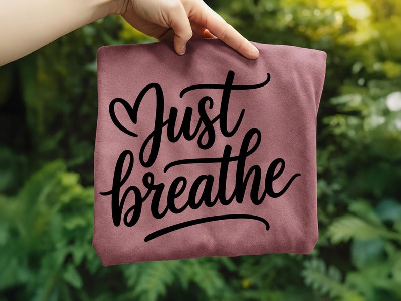 May include: A maroon-colored t-shirt with the words "Just breathe" in a black, cursive font. The word "Just" has a heart symbol above it. The shirt is held up against a blurred green background.