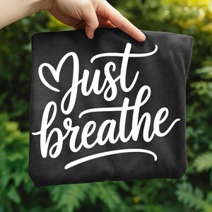May include: A black tote bag with the words "Just breathe" in white cursive script. The bag is held up against a blurred green background, suggesting an outdoor setting.
