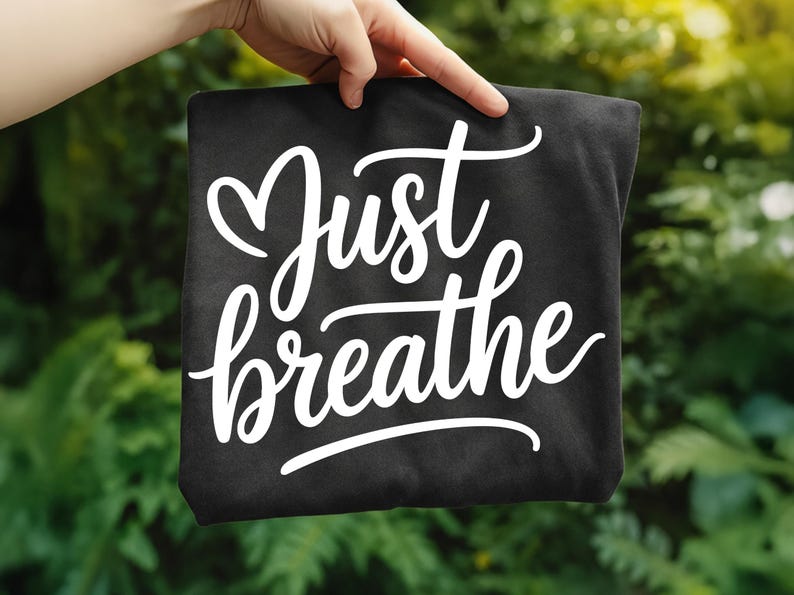 May include: A black tote bag with the words "Just breathe" in white cursive script. The bag is held up against a blurred green background, suggesting an outdoor setting.