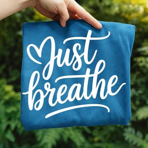 May include: A teal-colored fabric item with the words "Just breathe" in a white, cursive font. The word "Just" has a heart-shaped flourish above it. The item is held up against a blurred green background.