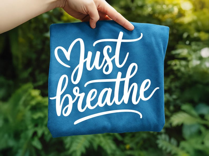 May include: A teal-colored fabric item with the words "Just breathe" in a white, cursive font. The word "Just" has a heart-shaped flourish above it. The item is held up against a blurred green background.