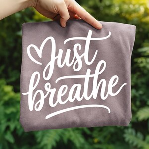 May include: A folded, dusty rose-colored t-shirt with the words "Just breathe" in a white, cursive font. The shirt is held up against a blurred green background of foliage.