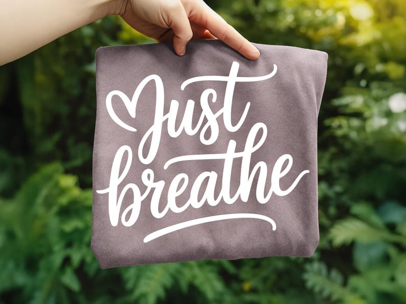May include: A folded, dusty rose-colored t-shirt with the words "Just breathe" in a white, cursive font. The shirt is held up against a blurred green background of foliage.