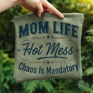 May include: A sage green pillow with the words "MOM LIFE Hot Mess Chaos Is Mandatory" in navy blue script. The pillow is held up by a hand against a blurred green background.