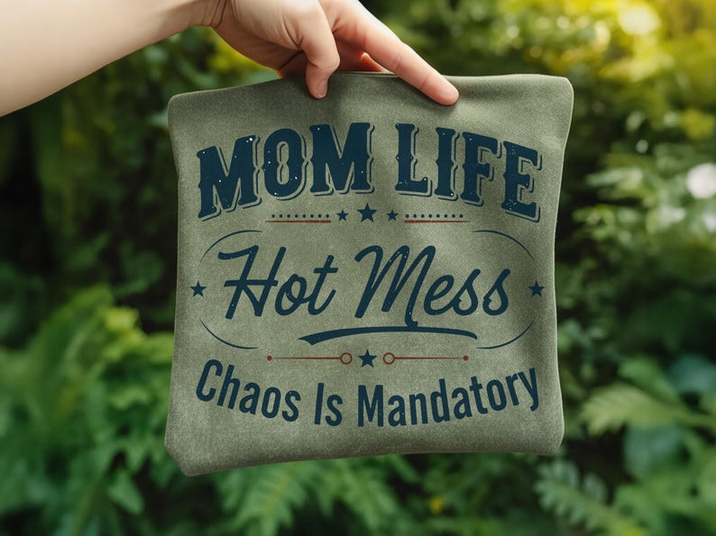 May include: A sage green pillow with the words "MOM LIFE Hot Mess Chaos Is Mandatory" in navy blue script. The pillow is held up by a hand against a blurred green background.