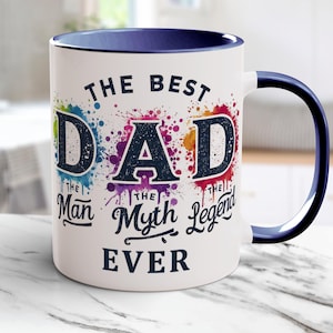 Op de afbeelding: Witte keramische mok met een blauw interieur en handvat. De mok heeft de tekst "THE BEST DAD EVER" in een kleurrijk verfspatontwerp. De woorden "The Man, The Myth, The Legend" zijn ook opgenomen.