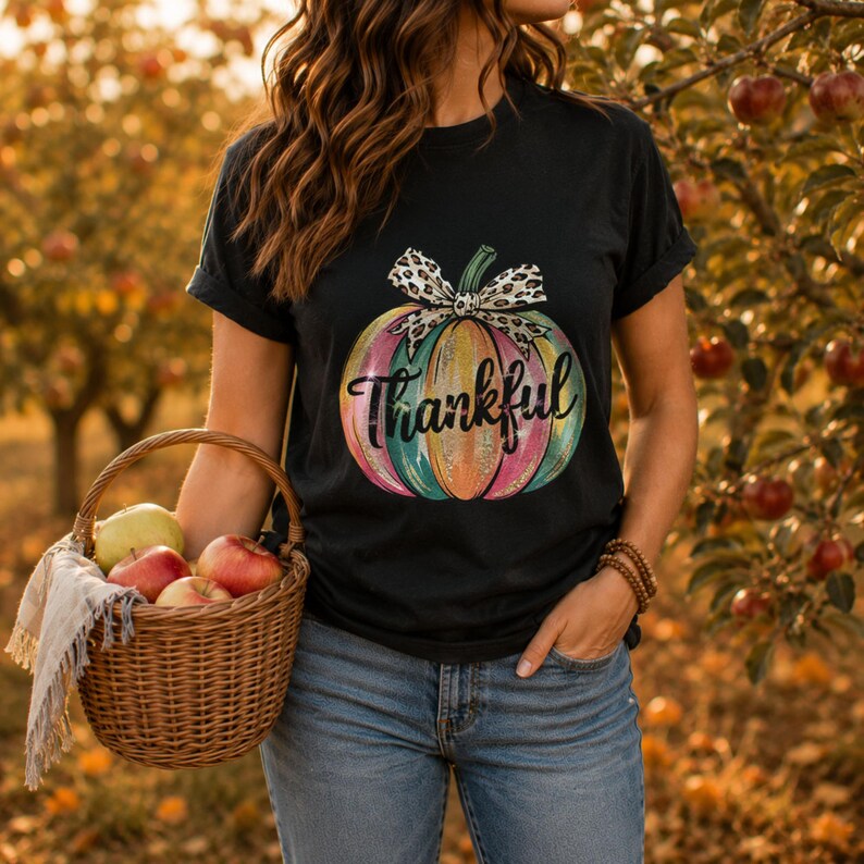 Woman wearing a black t-shirt featuring a colorful Thankful pumpkin graphic while picking fresh apples in an autumn orchard.