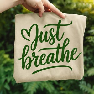 May include: A beige tote bag with the words "Just breathe" in green cursive script. The word "Just" is above "breathe", with a heart-shaped flourish above the "J". The background is a blurred green foliage.