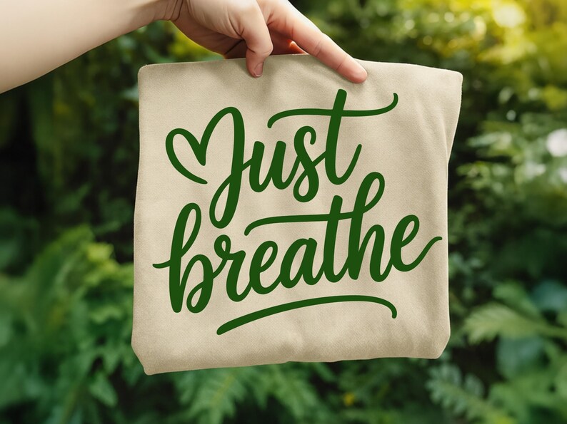 May include: A beige tote bag with the words "Just breathe" in green cursive script. The word "Just" is above "breathe", with a heart-shaped flourish above the "J". The background is a blurred green foliage.