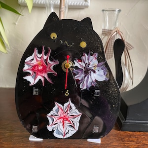 May include: A black cat-shaped clock with a whimsical design. The clock features a black face with gold accents and three abstract floral designs in shades of red, white, and purple. The clock hands are red and gold.