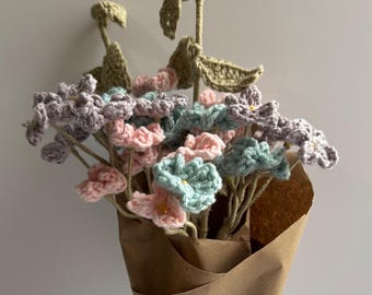 May include: A bouquet of crocheted flowers in shades of pink, purple, and blue, wrapped in brown paper and tied with a white lace ribbon.