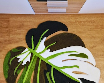Op de afbeelding: Een groen, wit en zwart tapijt in de vorm van een monstera-blad. Het tapijt is gemaakt van zacht, pluche materiaal en heeft een realistisch ontwerp.