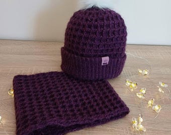 May include: A knitted plum-colored hat with a white pom-pom and matching neck warmer. The set has a textured waffle-like pattern. A small label is sewn on the hat. Decorative bee lights are scattered around the set.