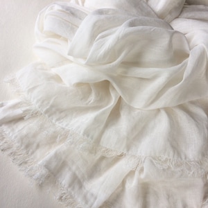 May include: A white scarf made of soft, flowing fabric. The scarf is draped and folded, with a fringed edge. The fabric appears lightweight and airy, suggesting a linen or similar material. The scarf is a solid, neutral color.