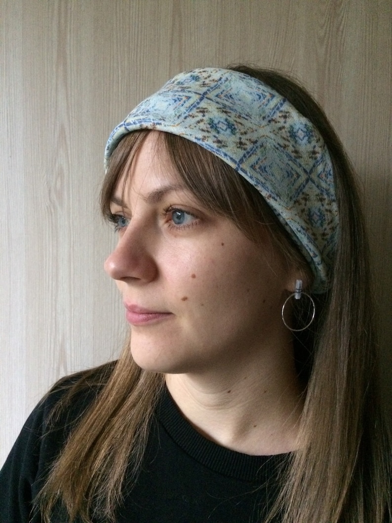 May include: A woman with long brown hair is wearing a blue and brown patterned headband. She is wearing a black shirt and has a silver hoop earring in her left ear.