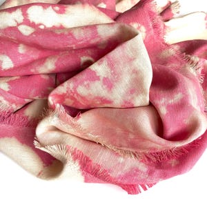 Może przedstawiać: Różowo-biały szalik z wzorem tie-dye. Szalik wykonany jest z miękkiej tkaniny i ma postrzępione krawędzie. Złożony i udrapowany, prezentuje wzór i fakturę. Modny dodatek.